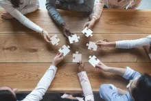 Photo of multiple hands in a circle holding puzzle pieces towards the centre of a table
